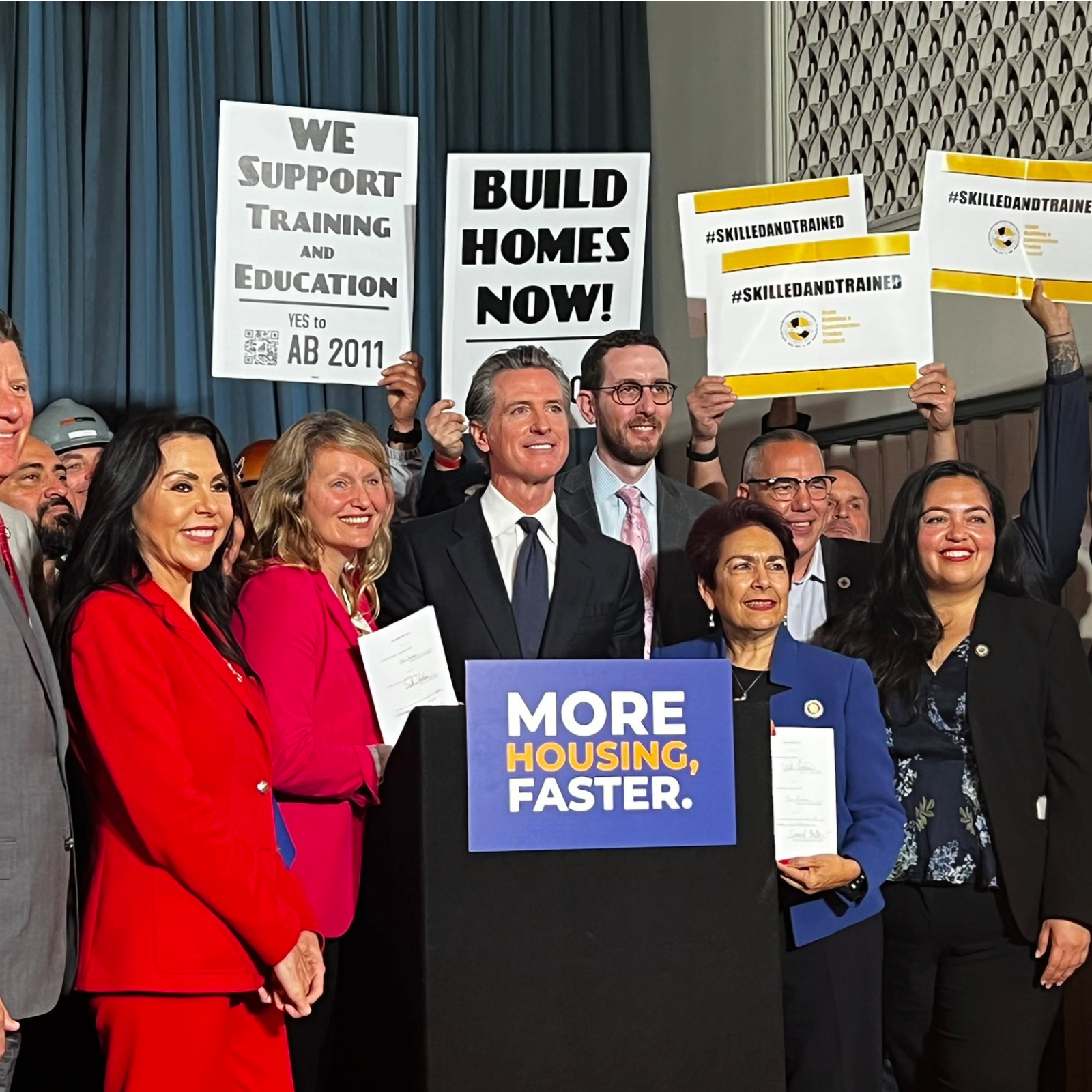 Legislators and construction workers around a podium with a sign saying 'more homes, faster'