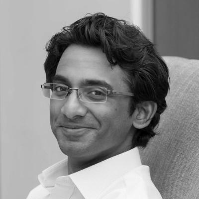 Smiling individual with glasses and dark hair, wearing a light-colored shirt, seated and looking towards the camera. Black and white image.