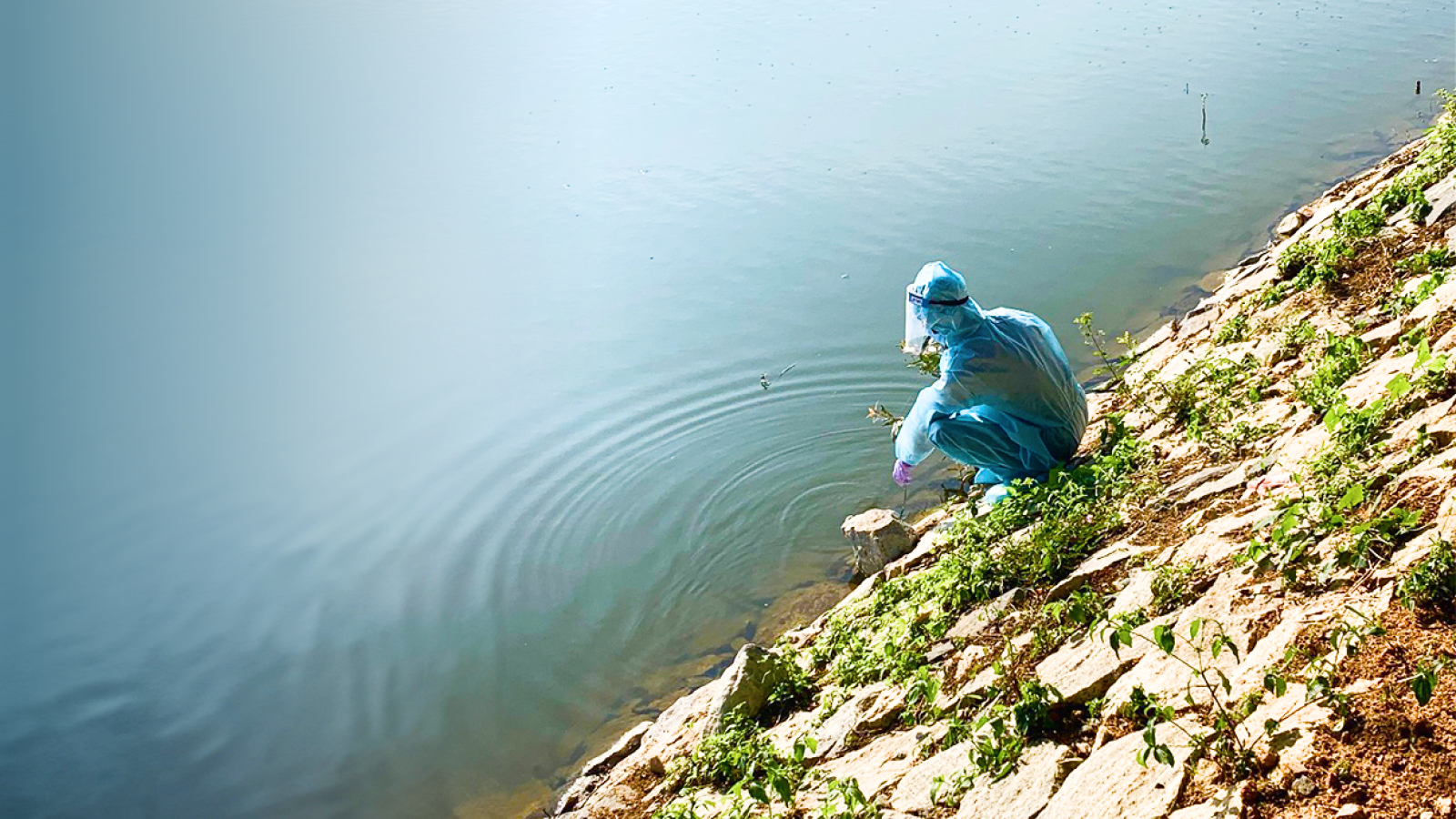 a woman in blue medical gear sitting on the edge of a lake.