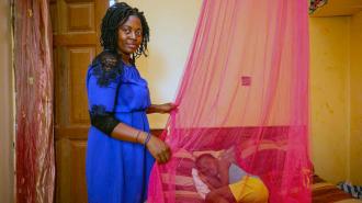 A woman in a blue dress standing next to a bed with a mosquito net. A child is on the bed.