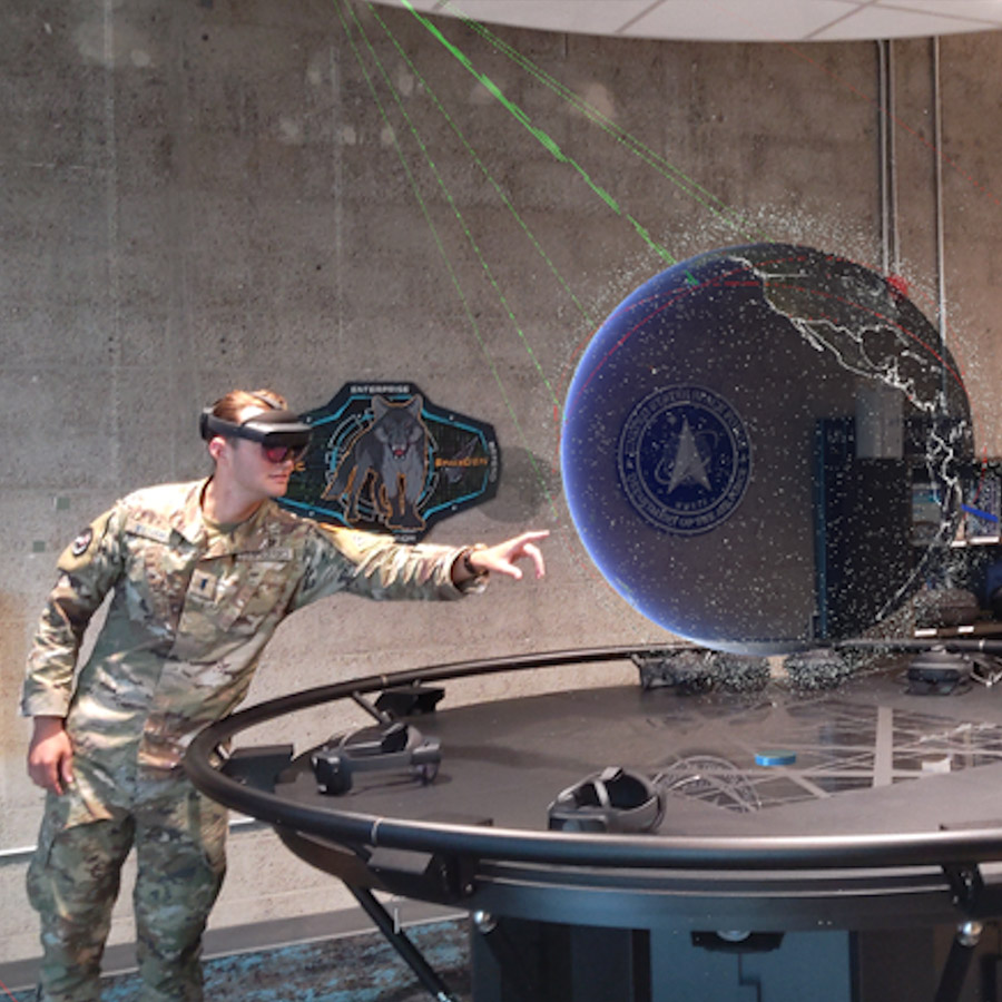 A man in camouflage is standing in front of a table with a virtual globe displayed above its surface