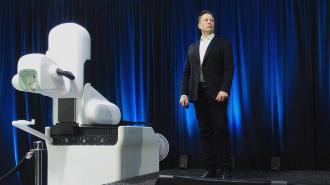 A man in a suit stands next to a robotic arm with computer-brain interfaces on a stage under blue lighting.