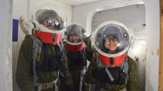 Three people in astronaut training suits and helmets smiling inside a spacecraft simulation chamber.