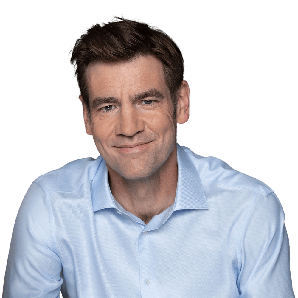 A man wearing a light blue shirt is smiling while looking directly at the camera.