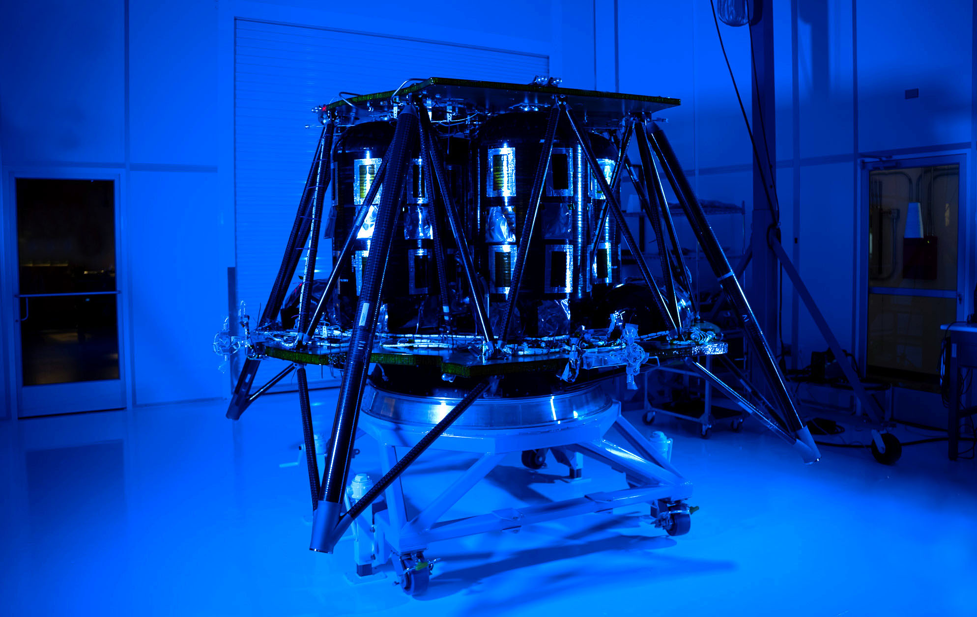 A fully assembled, illuminated spacecraft stands in a sterile room with blue lighting, showcasing its intricate components and structure.