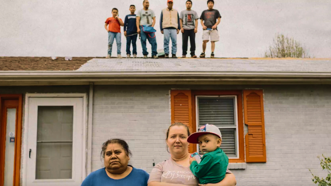Two women and a young boy stand in front of a house while five individuals stand on the roof in the background.