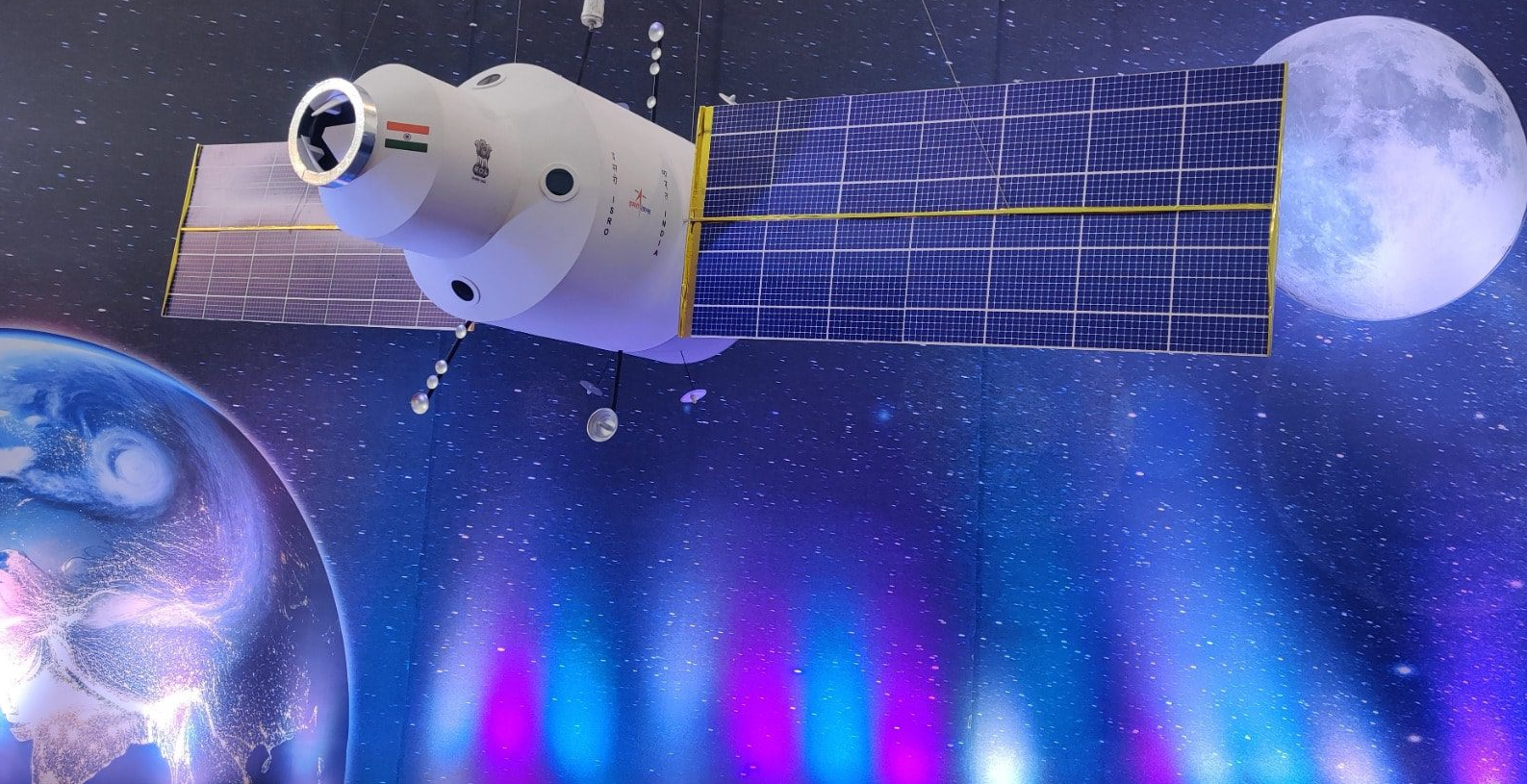 A space station model with solar panels floats in space, showcasing an Indian flag on its body. Earth and the Moon are visible in the background against a starry backdrop.