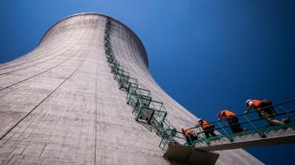 Three workers in orange vests and hard hats climb a ladder on the side of a massive concrete cooling tower of a nuclear power plant, against a clear blue sky.