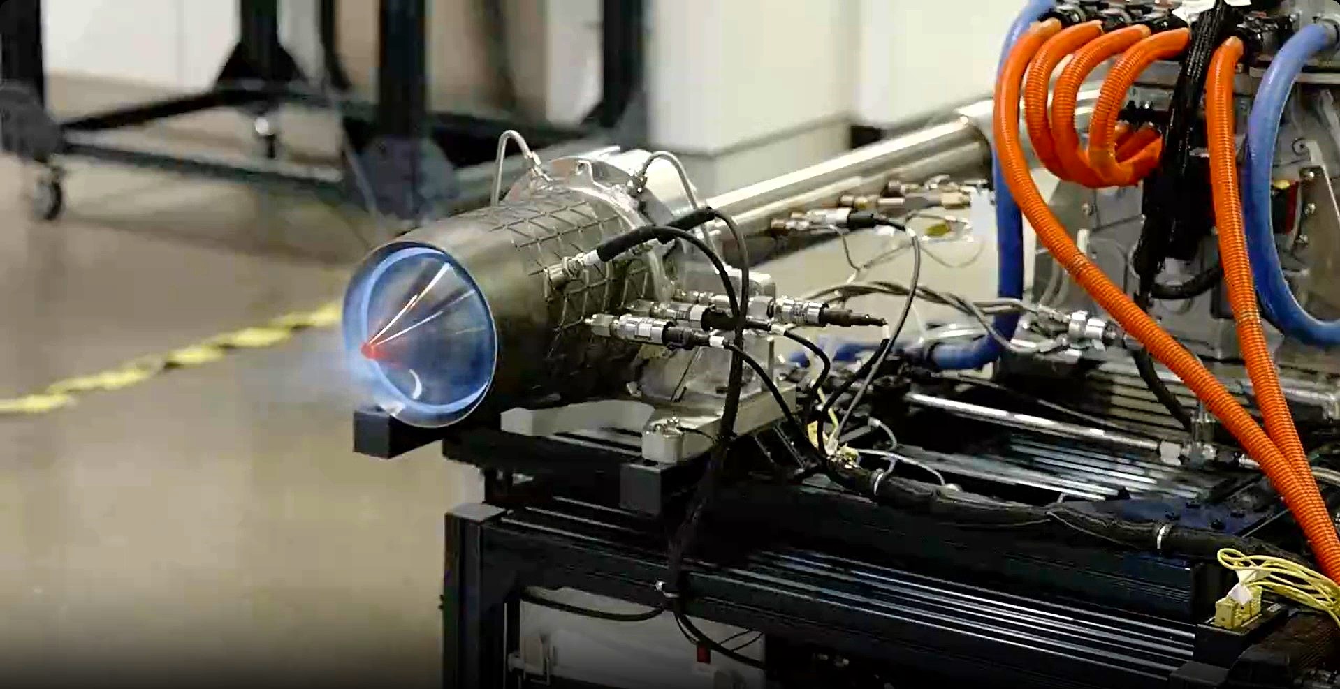 A close-up of an engine test firing, showcasing various connected hoses and cables, with the engine emitting blue flames from the nozzle, reminiscent of a powerful jet engine.