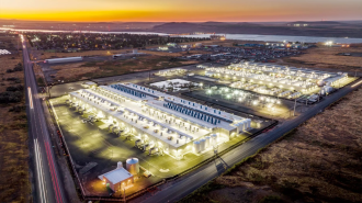 An aerial view of an illuminated industrial facility at dusk reveals multiple buildings, storage tanks, and surrounding roads. Nestled among fields with a distant waterway in the background, the scene evokes the intricate network of data centers operating silently beneath the serene landscape.