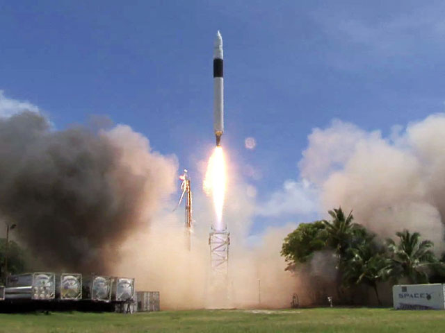 A rocket launches into the sky, surrounded by smoke, against a clear blue background with clouds. Palm trees and a clear sky are visible in the background.