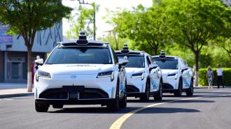 Three robotaxis glide in a line on a sunny road with trees and buildings in the background.