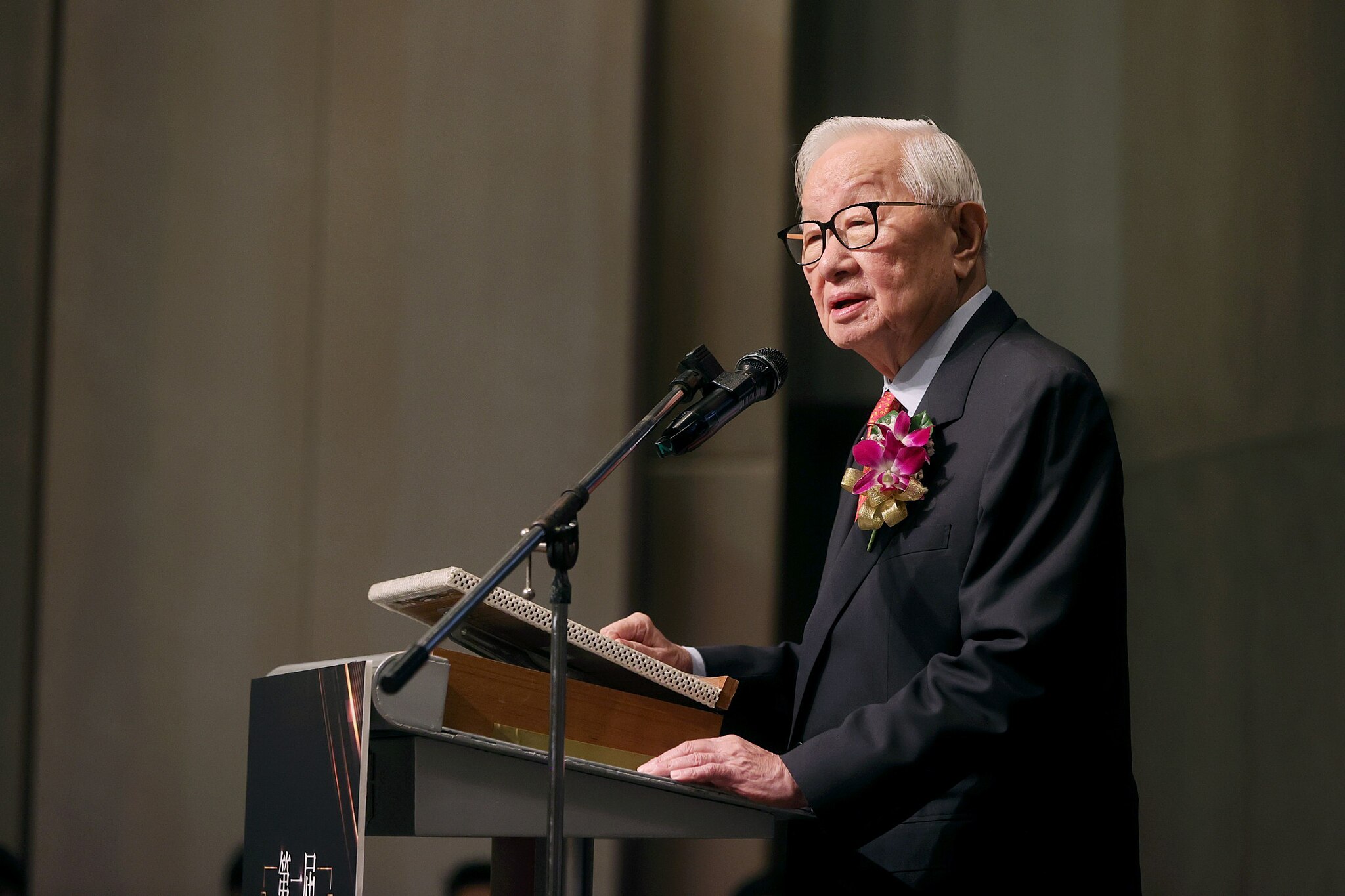 An elderly man in a suit speaks at a podium with a microphone, wearing glasses and a flower boutonniere.