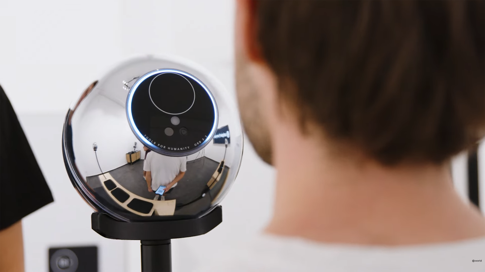 A silver spherical device on a stand, complete with a camera lens capturing a person. Reflections reveal the room's walls and furniture, lending an air of mystery akin to safeguarding one's personhood credentials.