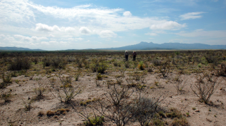 Two people trek across a dry, open desert landscape marked by sparse vegetation under a partly cloudy sky, illustrating the challenges of water scarcity. In the distance, mountains stand as silent observers to this arid expanse.