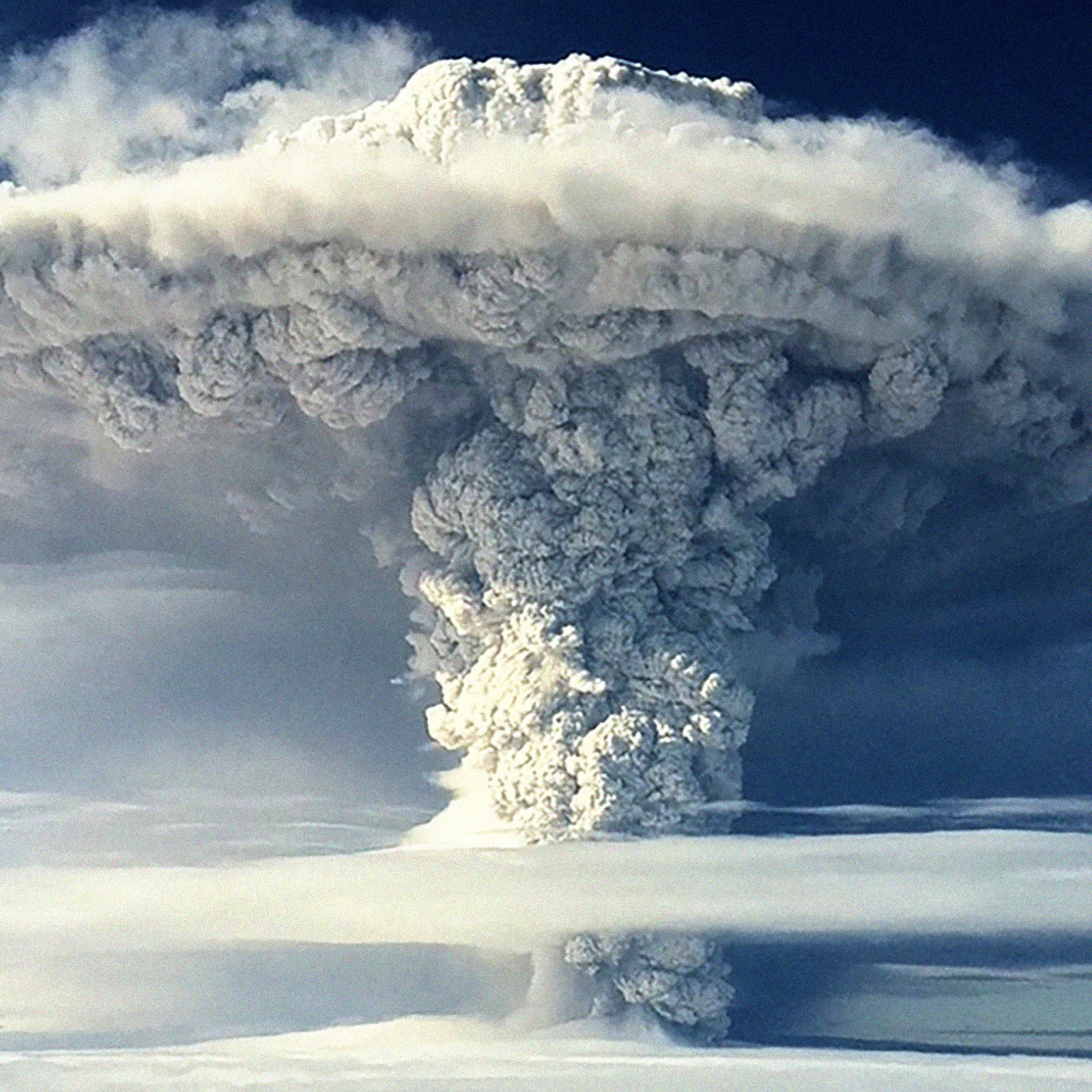 Massive plume of ash and smoke rises from a volcanic eruption, billowing into the sky with a mushroom-cloud shape against a blue background.