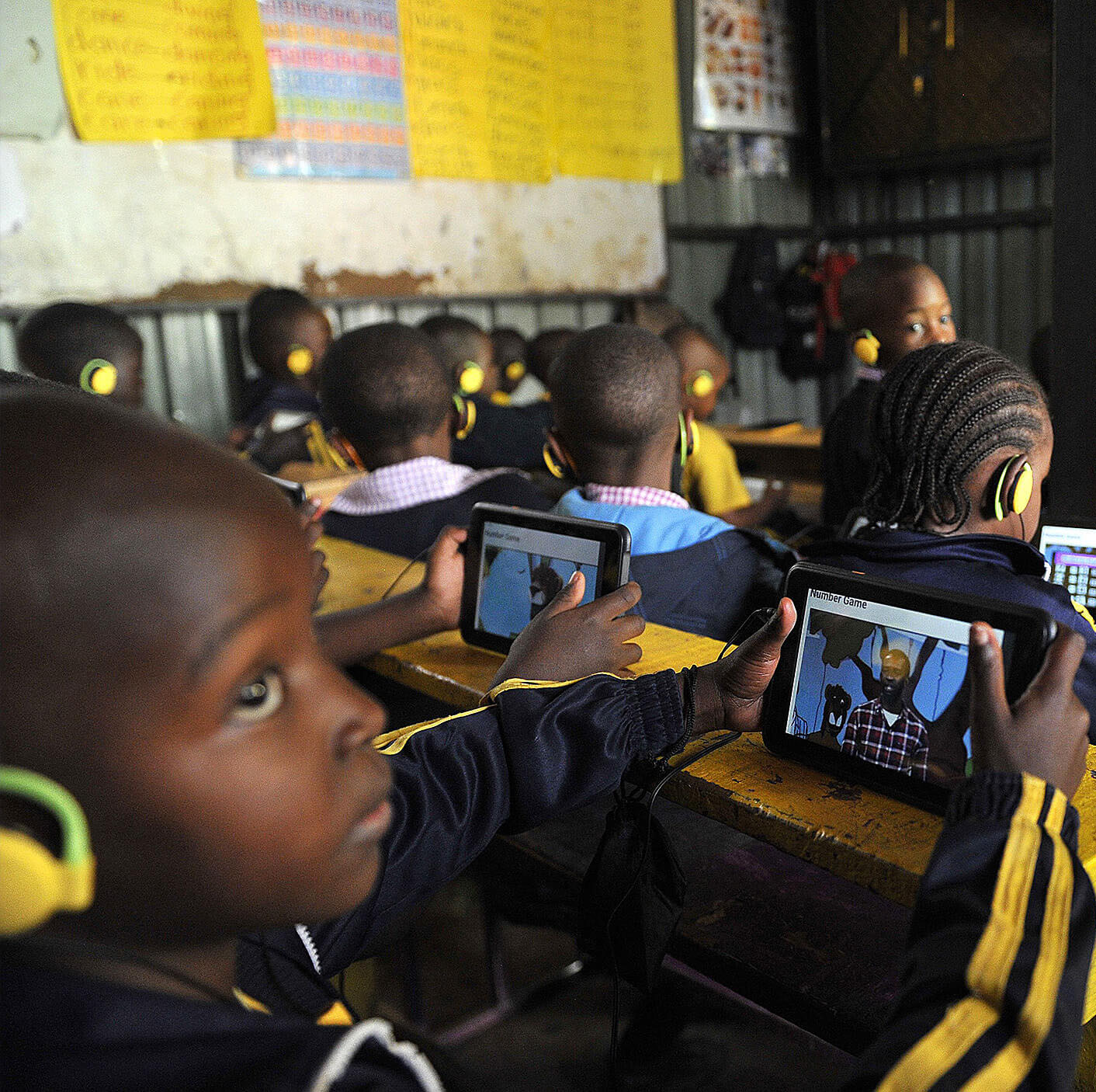 Children in a classroom use headphones and tablets at their desks, engaging with digital content.