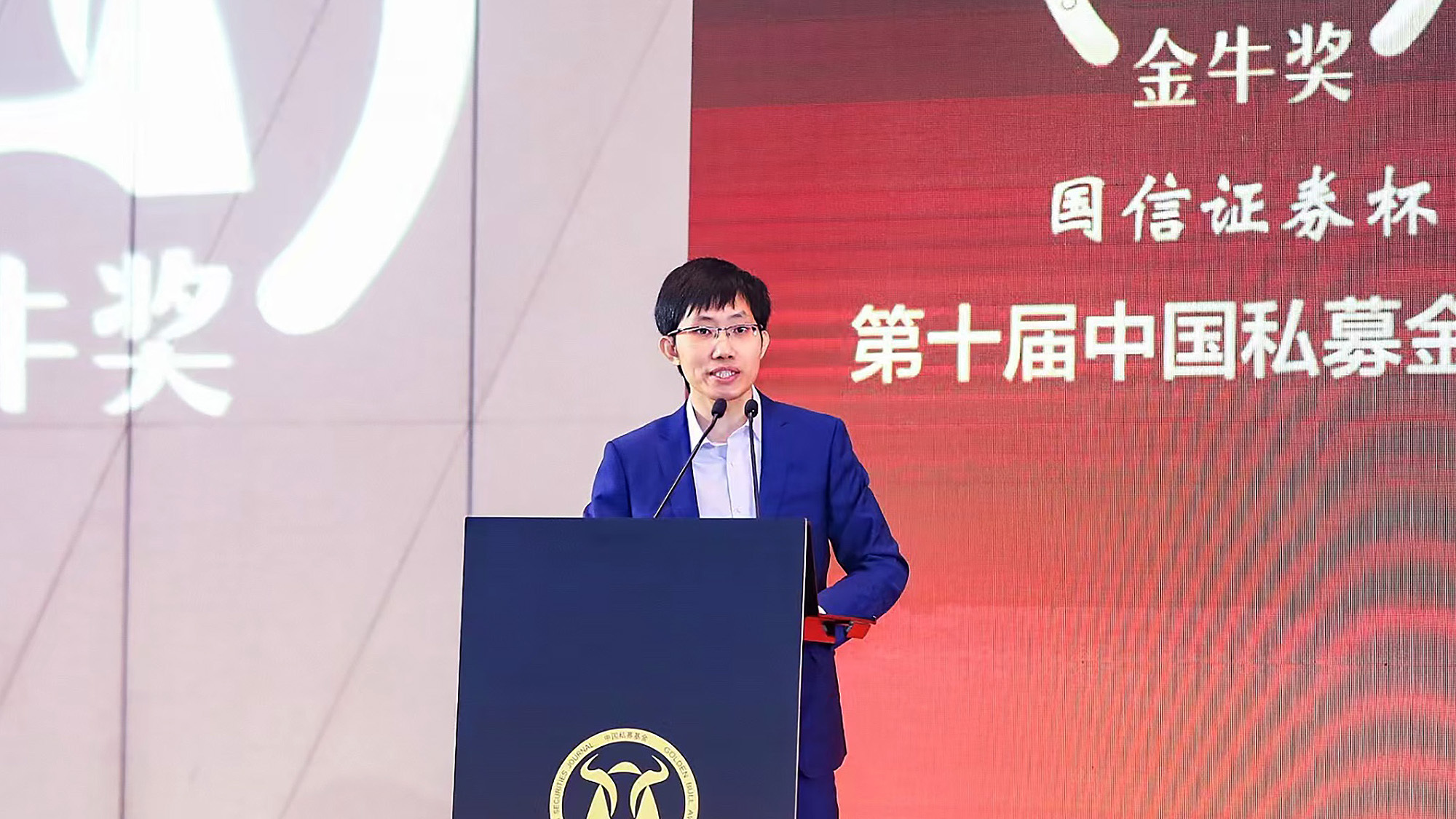 A person in a blue suit speaks at a podium during an event, with a large display screen in the background featuring Chinese text.