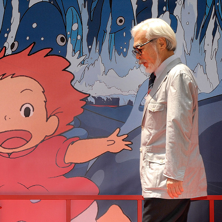 A person in a light jacket stands beside a large illustrated backdrop of a lively Studio Ghibli character.